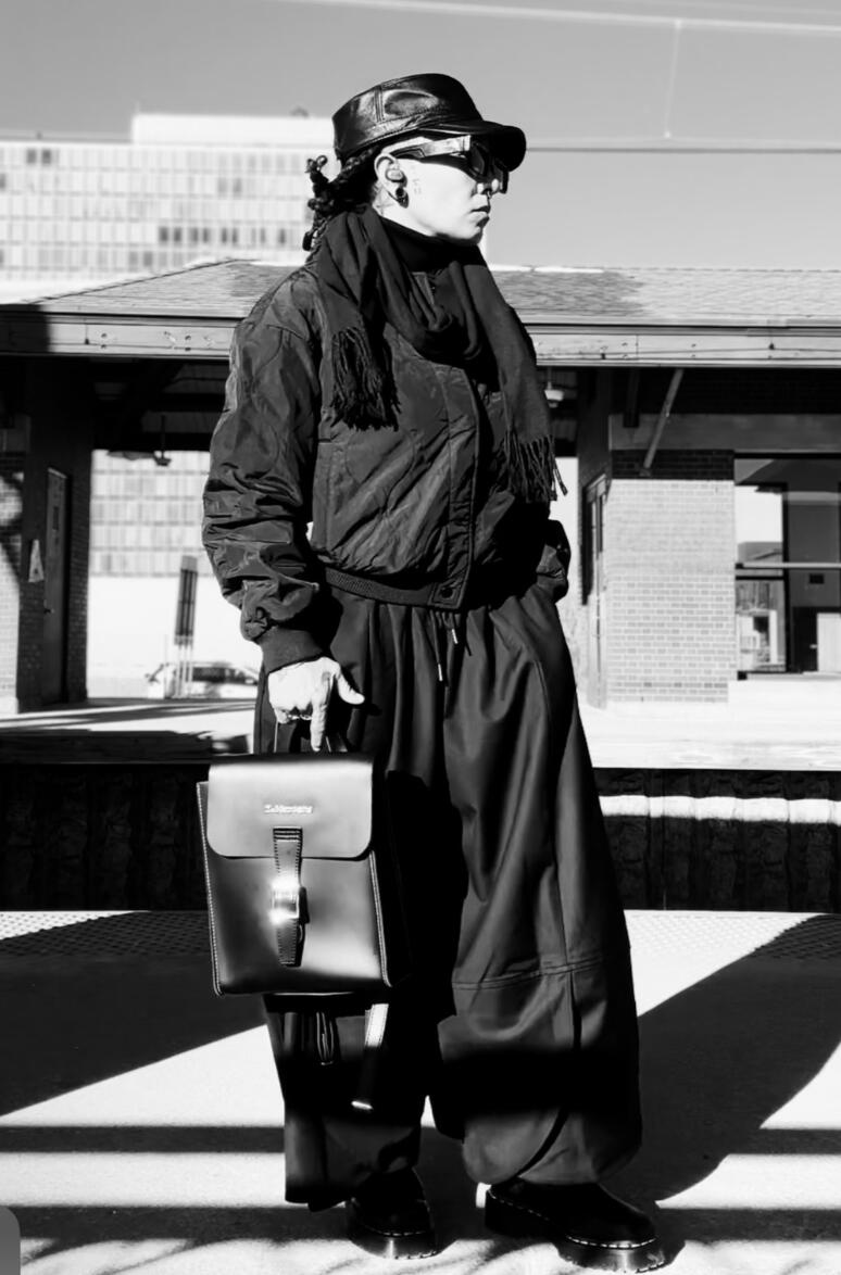 Black and white photo of Hannah standing at an outdoor train platform wearing all black, large sunglasses, and black hat, and looking off into the distance.